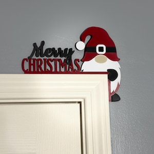 Close-up shot of a Merry Christmas gnome door corner decoration mounted on a white door frame against a grey wall.