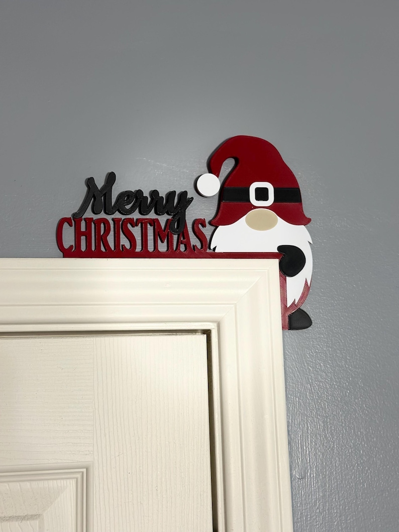 Close-up shot of a Merry Christmas gnome door corner decoration mounted on a white door frame against a grey wall.