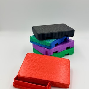 May include: A stack of colorful, rectangular storage boxes. The boxes are in various colors, including red, black, green, blue, and purple. The red box is open, revealing a small tray. The boxes have a textured surface.