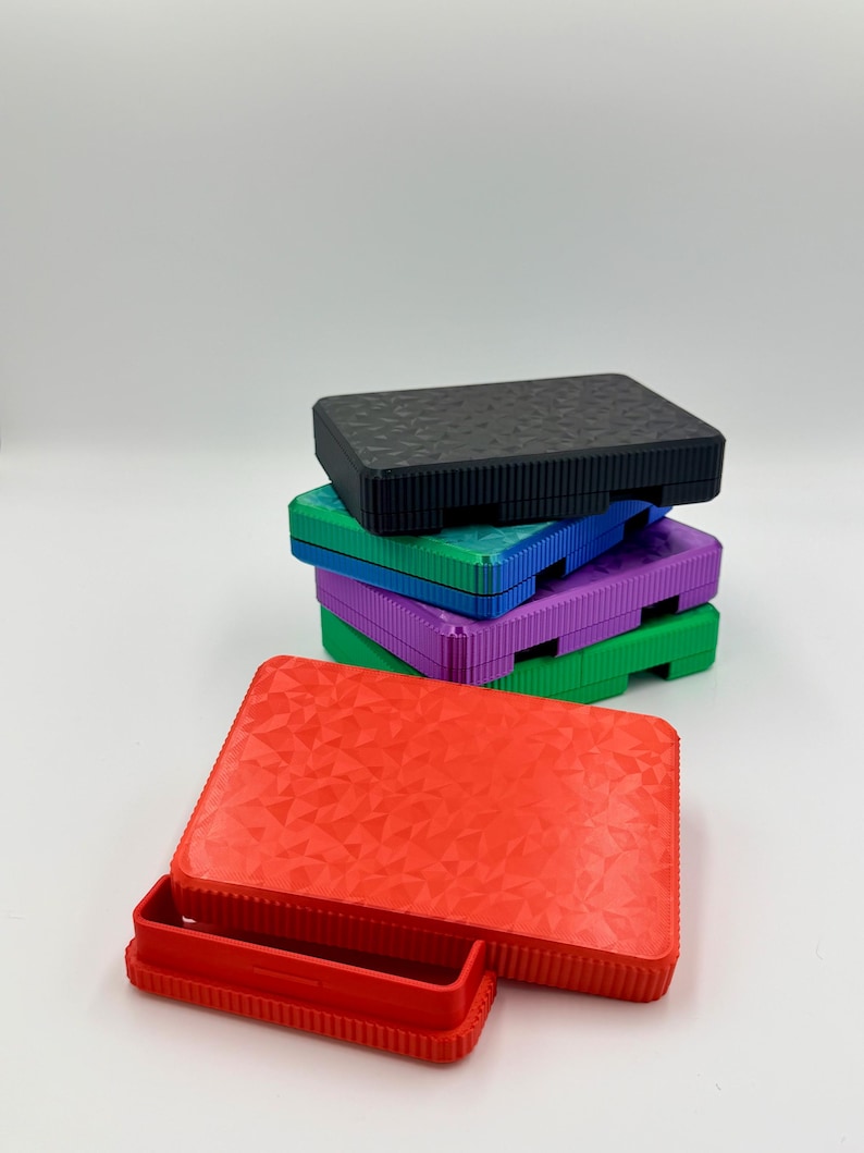 May include: A stack of colorful, rectangular storage boxes. The boxes are in various colors, including red, black, green, blue, and purple. The red box is open, revealing a small tray. The boxes have a textured surface.