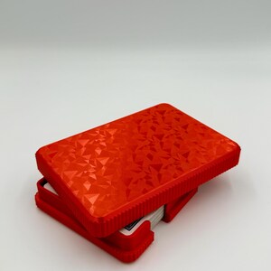 May include: A red playing card case with a geometric pattern on the lid. The case is open, revealing a deck of cards. The case is made of a hard plastic material and has a textured surface.