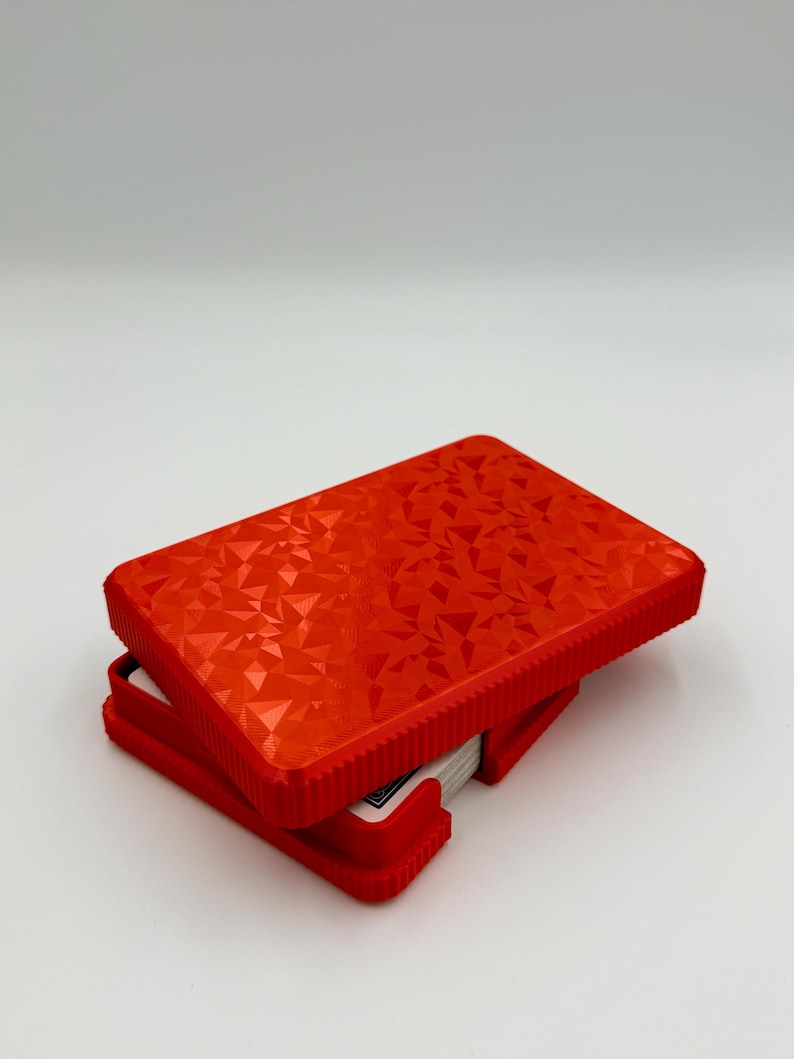 May include: A red playing card case with a geometric pattern on the lid. The case is open, revealing a deck of cards. The case is made of a hard plastic material and has a textured surface.
