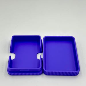 May include: A purple, rectangular box with a matching lid. The box is open, revealing the interior. The lid has a unique shape with two curved cutouts. The box and lid have a ridged texture around the edges.