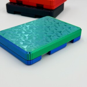May include: Three rectangular boxes in red, black, and teal. The teal box has a geometric pattern on the lid and is stacked on a blue base. The red and black boxes are stacked behind the teal box. The boxes have a ridged design on the sides.