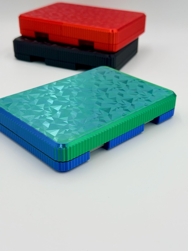 May include: Three rectangular boxes in red, black, and teal. The teal box has a geometric pattern on the lid and is stacked on a blue base. The red and black boxes are stacked behind the teal box. The boxes have a ridged design on the sides.
