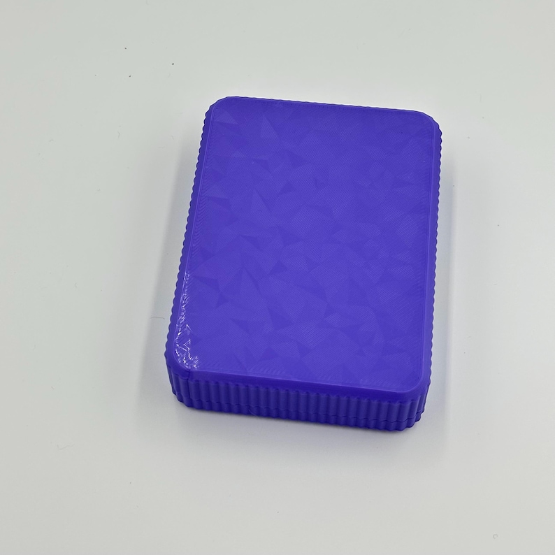 May include: A rectangular, solid purple box with a textured, geometric pattern on the top surface. The edges of the box have a ribbed design. The box is sitting on a white surface.