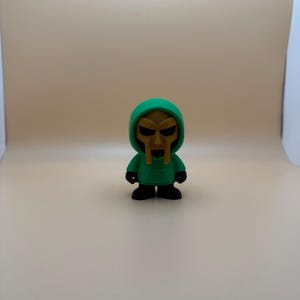 May include: A small figurine of a character wearing a green hooded sweatshirt and a gold mask. The figure has black shoes and a gold face mask with a stern expression. The figurine is standing on a neutral background.