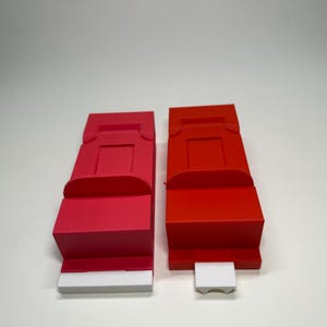 以下が含まれることがあります： 白い土台に、赤い収納容器とピンクの収納容器が2つあります。容器は長方形で、上部に小さな開口部があります。