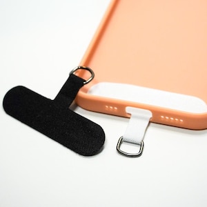 Peut inclure: Une dragonne de téléphone noire et blanche avec un anneau métallique. La dragonne est attachée à une coque de téléphone couleur pêche.