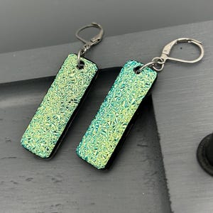 May include: A pair of rectangular earrings with a textured, iridescent green surface. The earrings have a black border and silver-colored lever-back closures. The earrings are displayed on a dark gray surface.