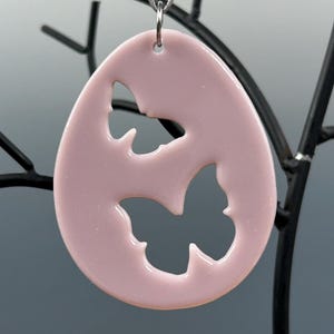 Op de afbeelding: Een roze keramische eivormige hanger met twee vlinderuitsnijdingen. De hanger hangt aan een zilveren koord.