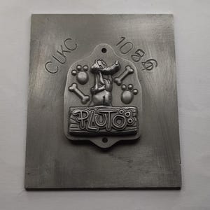 Könnte beinhalten: Eine silberne Metallform mit dem Text "CLKC 1086" und einem erhabenen Bild von Pluto dem Hund mit dem Text "PLUTO's" auf einem Banner.