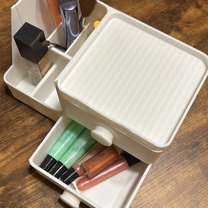 May include: A white plastic makeup organiser featuring a tiered design with open compartments for brushes and a square bottle, plus a pull-out drawer containing several lip gloss tubes, including green and copper-toned cosmetic products.