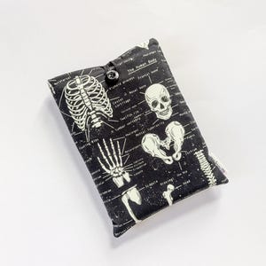 May include: Black and white padded case featuring a skeletal diagram of the human body. The case has a button closure and includes labeled illustrations of bones and anatomical structures. The text "The Human Body" is visible.