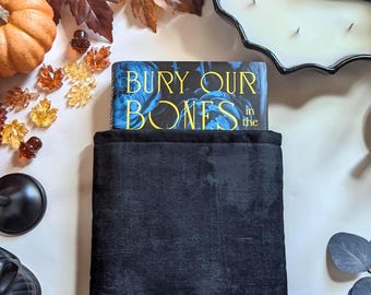 May include: A book titled "Bury Our Bones" with a blue and gold cover rests on a black blanket. The scene is decorated with fall-themed elements, including a pumpkin, decorative leaves, and a bat-shaped candle.
