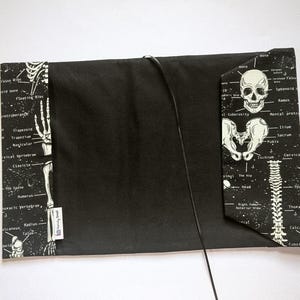 May include: Black book cover with a skeletal anatomy design. The cover features a black background with white illustrations of bones and anatomical labels. A black cord is attached to the cover. The cover has a rectangular shape.