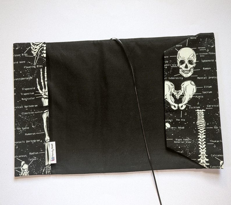 May include: Black book cover with a skeletal anatomy design. The cover features a black background with white illustrations of bones and anatomical labels. A black cord is attached to the cover. The cover has a rectangular shape.