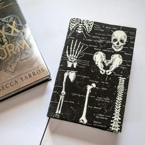May include: A book cover with the title "Iron Flame" and author "Rebecca Yarros" next to a black book cover with a white skeletal diagram. The diagram includes bones, labels, and a skull.