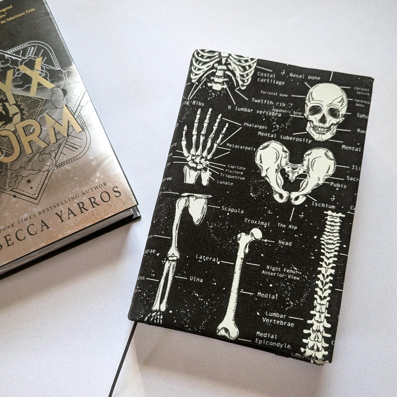 May include: A book cover with the title "Iron Flame" and author "Rebecca Yarros" next to a black book cover with a white skeletal diagram. The diagram includes bones, labels, and a skull.