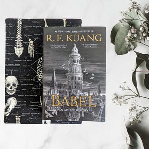 May include: A book titled "BABEL An Arcane History" by R.F. Kuang, a New York Times bestseller, is displayed next to a black and white book sleeve. The book cover features a detailed illustration of a tall building and cityscape.