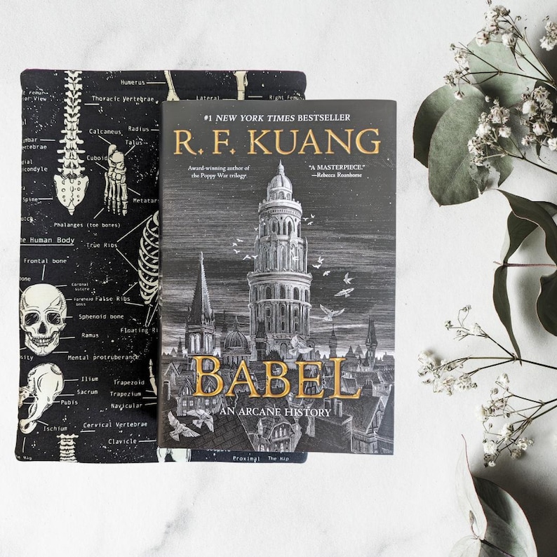 May include: A book titled "BABEL An Arcane History" by R.F. Kuang, a New York Times bestseller, is displayed next to a black and white book sleeve. The book cover features a detailed illustration of a tall building and cityscape.