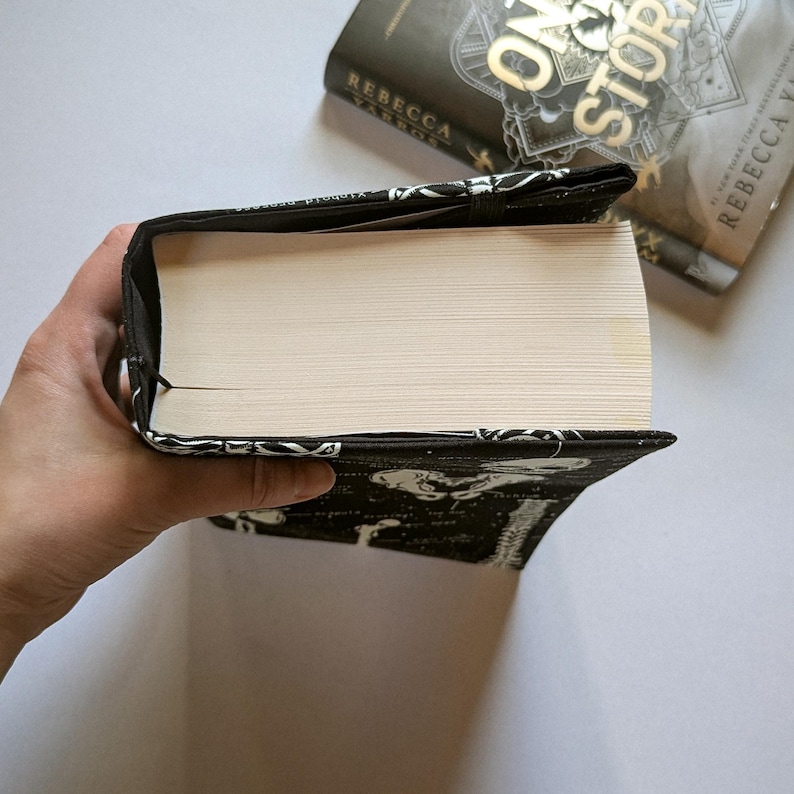May include: A black and white book cover with a patterned design, protecting a book. The book cover is wrapped around a thick book with white pages. The book cover is made of fabric and has a black elastic band.