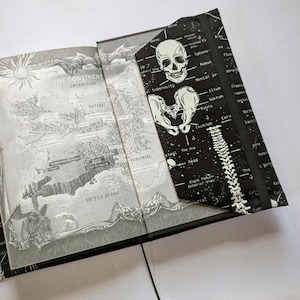 May include: Open journal with a map illustration and a black cover featuring a white skull and skeletal anatomy. The map shows a continent with the text "The Continent" and "Emerald Sea". The journal is open to a page with a detailed map.