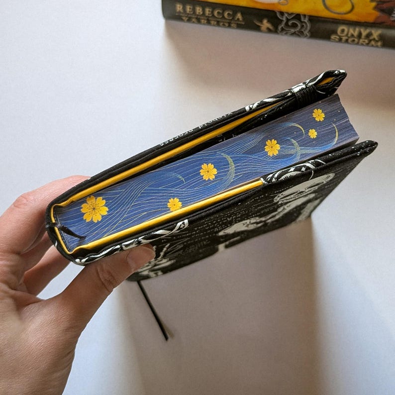 May include: A black book cover with a white design and yellow trim, open to reveal a blue page with yellow flower accents. A black bookmark is attached. The book cover is held by a hand.
