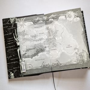 May include: Open book with a detailed map illustration in grayscale. The map features a continent with geographical features and place names. The left page has a black background with skeletal diagrams and anatomical labels.