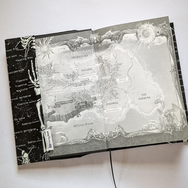 May include: Open book with a detailed map illustration in grayscale. The map features a continent with geographical features and place names. The left page has a black background with skeletal diagrams and anatomical labels.