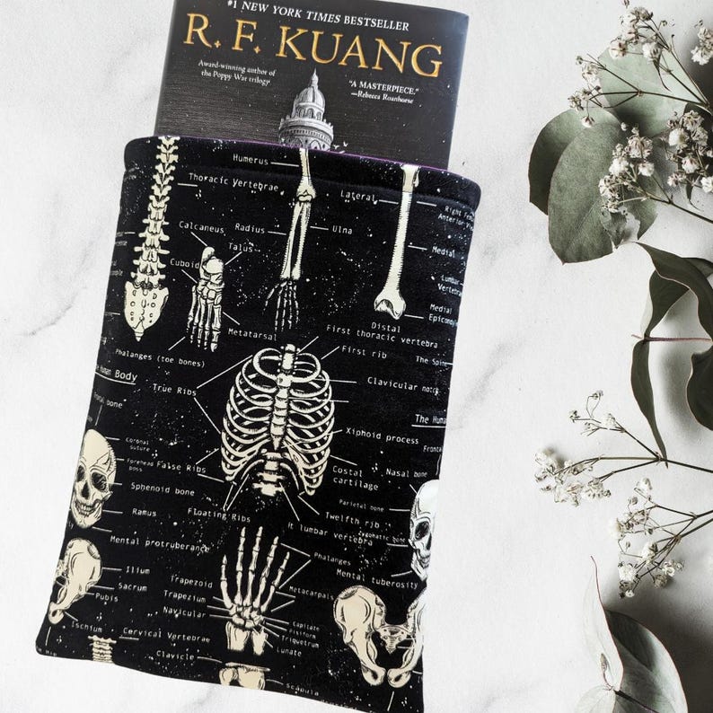 May include: Black book sleeve with a white skeletal diagram print. The anatomical design includes bones, labels, and text. A book with the title "R. F. Kuang" is partially visible inside the sleeve. The sleeve is designed to protect books.