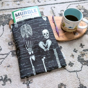 May include: A black book sleeve with a white skeletal diagram design, holding a book titled "MURDLE Volume 3". A mug of coffee and a pen rest on a wooden tray. The mug has the word "VIENNA" on it.