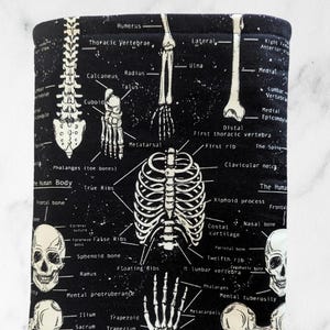 May include: Black and white anatomical print featuring detailed illustrations of the human skeletal system, including the skull, spine, ribs, and various bones. The text labels the different parts of the skeleton. The background is black.