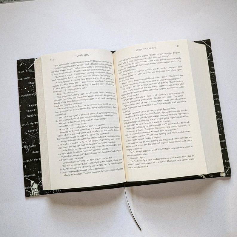 May include: An open book with black covers and a black bookmark. The pages are filled with printed text. The left cover has a black and white design with anatomical terms. The book is open to pages 148 and 149.