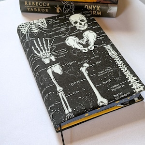 May include: A black book cover with a white skeletal diagram design. The cover features illustrations of bones, including a skull, ribs, and spine, with anatomical labels. The book has a yellow edge and a black ribbon bookmark.