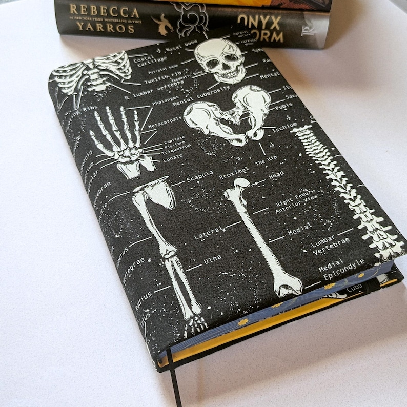 May include: A black book cover with a white skeletal diagram design. The cover features illustrations of bones, including a skull, ribs, and spine, with anatomical labels. The book has a yellow edge and a black ribbon bookmark.