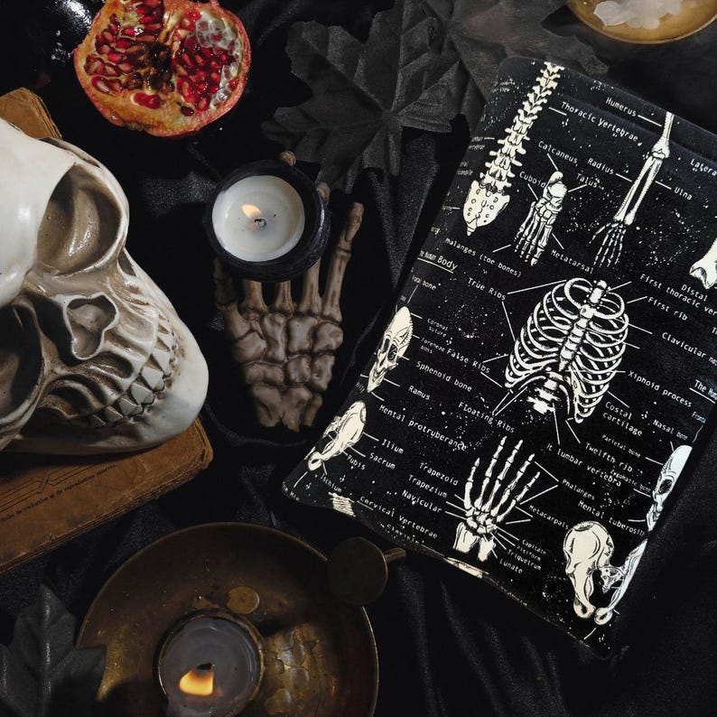 May include: A black and white anatomical pillow featuring skeletal diagrams, including the rib cage and hand bones. The image also includes a skull, a pomegranate, a candle, and a skeleton hand.