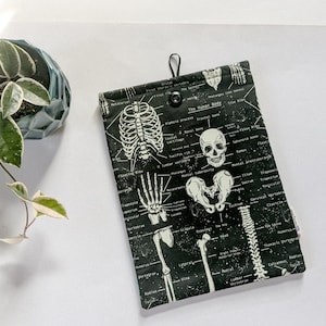 May include: Black and white anatomical print tablet sleeve featuring a detailed illustration of the human skeletal system. The sleeve has a black button closure and a loop for hanging. The text "The Human Body" is visible.