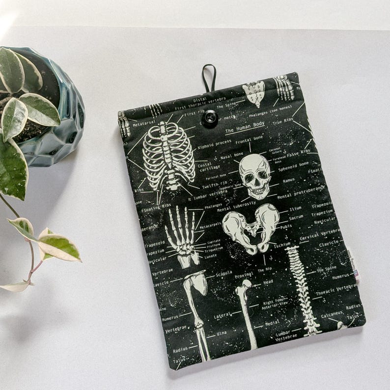 May include: Black and white anatomical print tablet sleeve featuring a detailed illustration of the human skeletal system. The sleeve has a black button closure and a loop for hanging. The text "The Human Body" is visible.