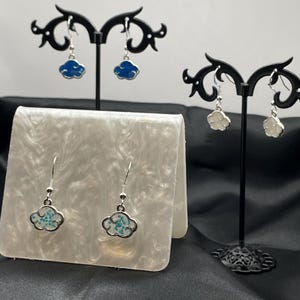 May include: A pair of silver dangle earrings with a cloud design. One pair features a blue cloud with white speckles, and the other pair features a white cloud with blue speckles.