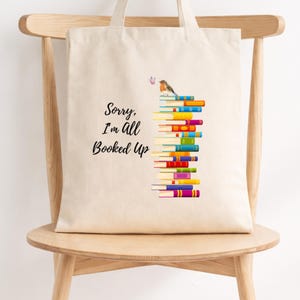 May include: A beige canvas tote bag featuring a stack of colourful books with a small bird perched on top. The text "Sorry, I'm All Booked Up" is printed in black script to the left of the book stack. The design includes various book spine colours.