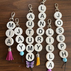 May include: Six personalized keychains featuring white circular beads with black letters spelling the names Mia, Jay, Autumn, Landry, Lilla, and Joseph. The keychains include silver lobster clasps, colorful tassels, butterfly charms, and silicone beads.