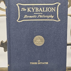 Puede incluir: Un libro azul marino titulado "The Kybalion" con letras doradas y un emblema dorado. El libro se sostiene en un atril de metal dorado. Tambi&eacute;n se ve el texto "Hermetic Philosophy" y "By Three Initiates".