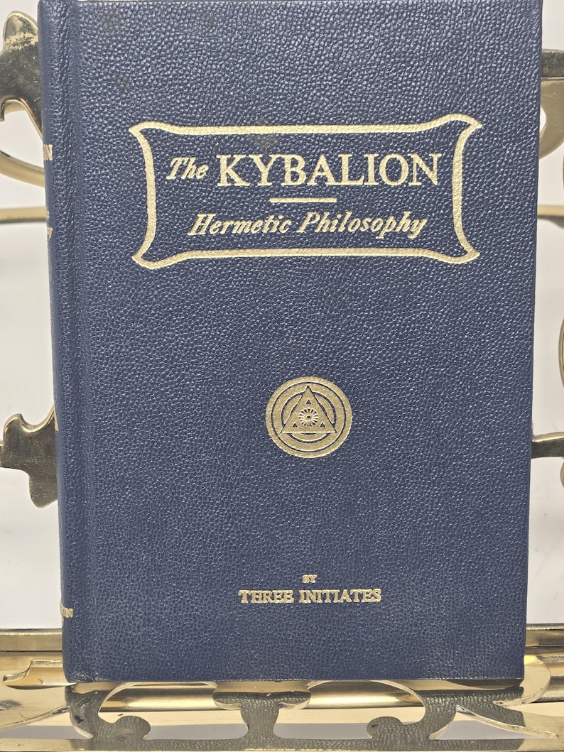 Puede incluir: Un libro azul marino titulado "The Kybalion" con letras doradas y un emblema dorado. El libro se sostiene en un atril de metal dorado. Tambi&eacute;n se ve el texto "Hermetic Philosophy" y "By Three Initiates".