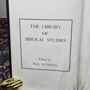 May include: An open book titled "The Library of Biblical Studies" with text in a black frame. The book is held open by a gold-colored hand-shaped book holder. The book's cover is dark blue.
