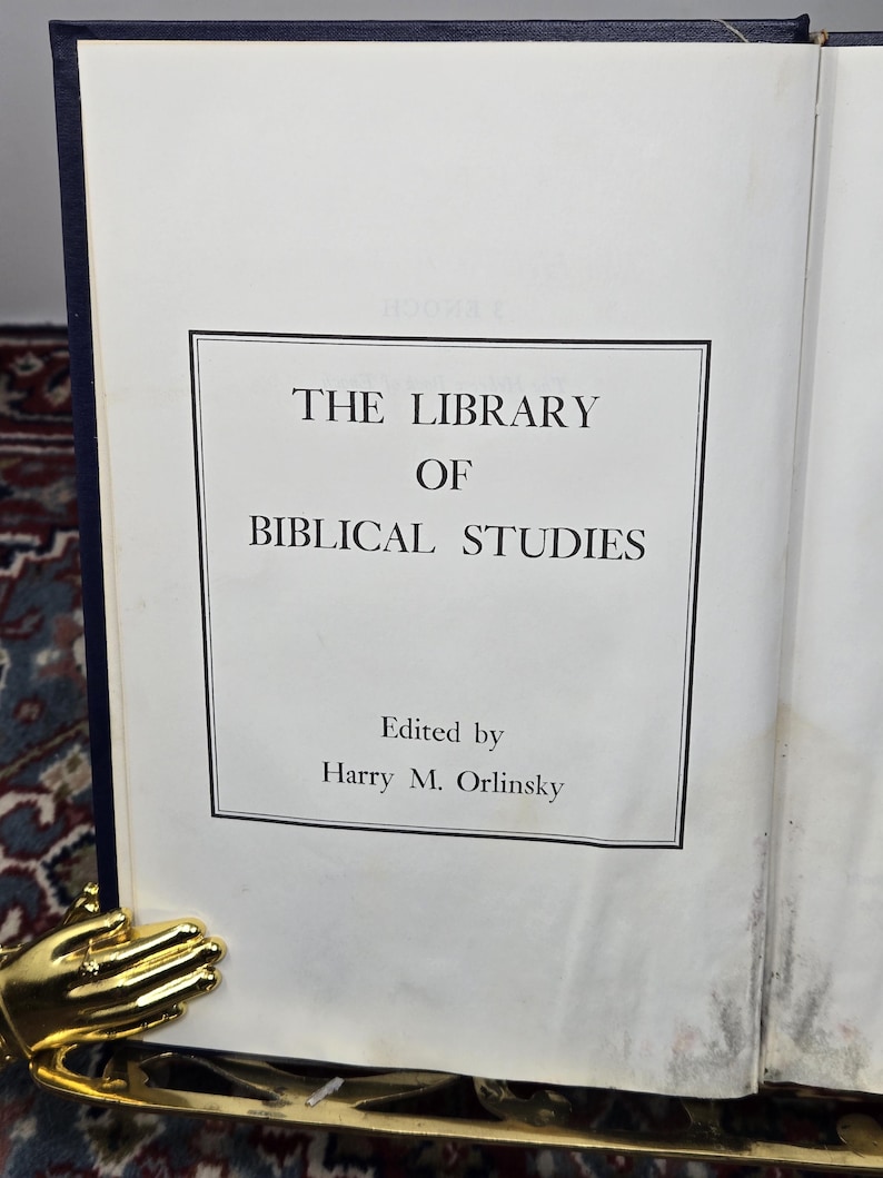 May include: An open book titled "The Library of Biblical Studies" with text in a black frame. The book is held open by a gold-colored hand-shaped book holder. The book's cover is dark blue.