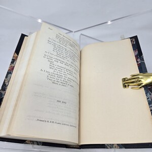 May include: An open book with a cream-colored interior and a black cover with a floral pattern. The book is open to the last page, which has the text "THE END" printed on it. The book is sitting on a table with a gold-colored hand-shaped paperweight.