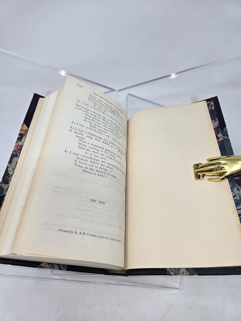 May include: An open book with a cream-colored interior and a black cover with a floral pattern. The book is open to the last page, which has the text "THE END" printed on it. The book is sitting on a table with a gold-colored hand-shaped paperweight.