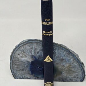 Puede incluir: Un libro azul marino titulado "The Kybalion: Hermetic Philosophy" con letras doradas, de pie en un sujetalibros de &aacute;gata azul y gris. El libro tiene un emblema dorado y es publicado por la Yogi Publication Society.