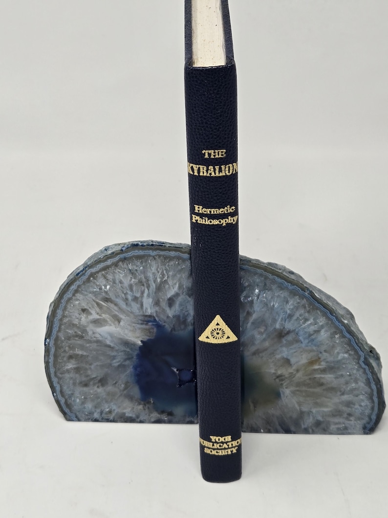 Puede incluir: Un libro azul marino titulado "The Kybalion: Hermetic Philosophy" con letras doradas, de pie en un sujetalibros de &aacute;gata azul y gris. El libro tiene un emblema dorado y es publicado por la Yogi Publication Society.